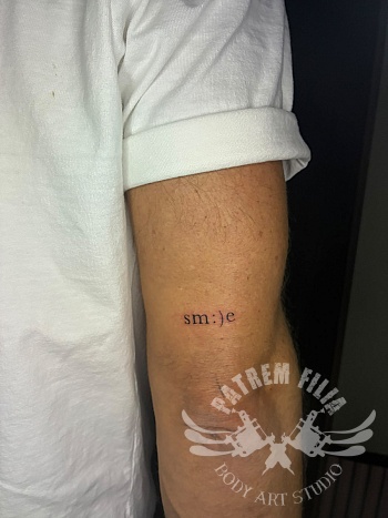 Minimalistische 'sm:)e' typografie tattoo op bovenarm